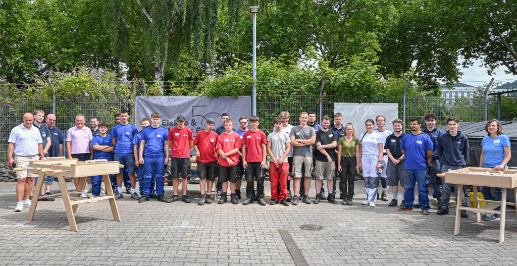 Gruppenfoto von etwa 25 Personen in Arbeitskleidung stehend auf einem gepflasterten Außengelände vor einem Metallzaun mit Büschen, zwei Wikingertischen aus Holz stehen vorne links und rechts im Bild.