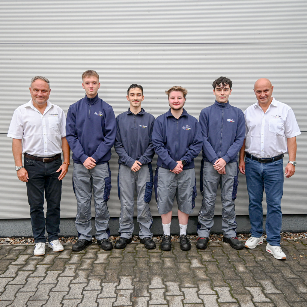 Gruppenfoto von sechs Männern vor einer weißen Wand, davon zwei ältere in weißem Hemd und vier jüngere in blauer Arbeitsjacke und grauer Arbeitshose mit kurzen Ärmeln, alle stehen auf gepflastertem Boden und schauen in die Kamera