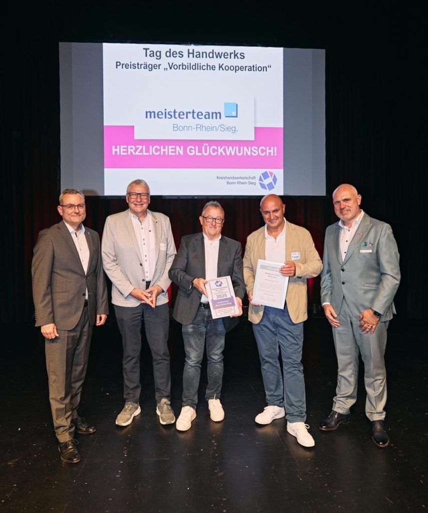 Fünf Männer in Anzügen stehen auf einer Bühne vor einer Leinwand mit der Aufschrift ‚Tag des Handwerks – Preisträger Vorbildliche Kooperation‘ und ‚meisterteam Bonn-Rhein/Sieg – Herzlichen Glückwunsch!‘, zwei von ihnen halten Urkunden.