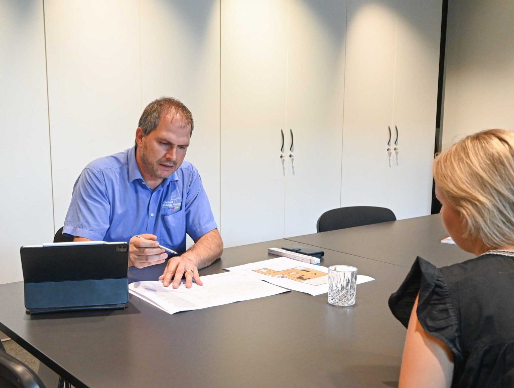 Ein Mann in blauem Hemd erklärt einer Frau mit blonden Haaren am Tisch Baupläne in einem modernen Büro mit weißen Schränken im Hintergrund.