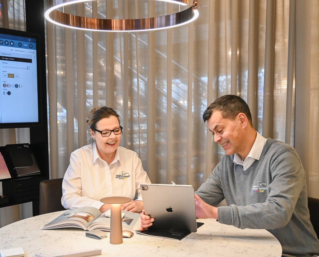 Zwei Menschen mit Firmenkleidung sitzen an einem runden Tisch mit Marmorplatte, schauen lachend auf ein geöffnetes Booklet und ein iPad, im Hintergrund Vorhänge und ein beleuchteter runder Deckenring.